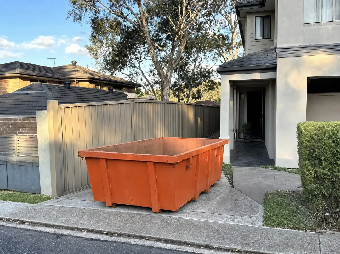An empty orange skip bin tucked lengthways into the narrow concrete driveway beside a two-storey Charlestown townhouse, a colorbond fence close on one side and a tall gum leaning over it