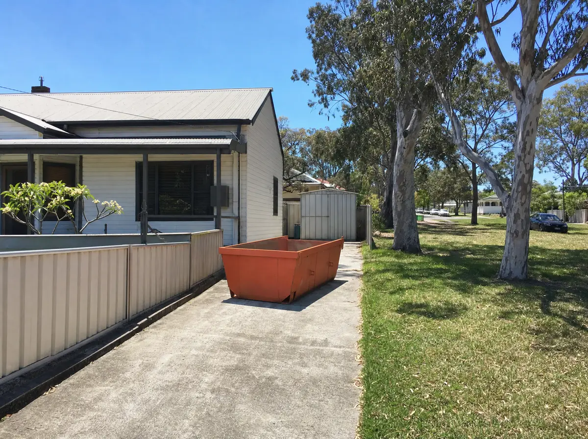 An empty orange skip bin sitting lengthways on the concrete driveway beside a white weatherboard miners cottage in Kurri Kurri, spotted gums along the wide grassed verge and a tin shed at the rear