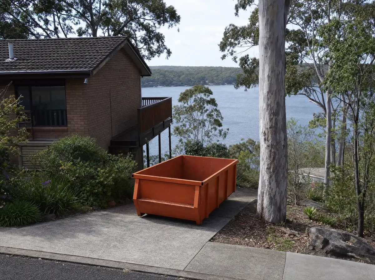 An empty orange skip bin sitting lengthways on the steep sloping driveway of a split-level Lake Macquarie home, gums and an angophora framing the view, the lake and its far shoreline visible below