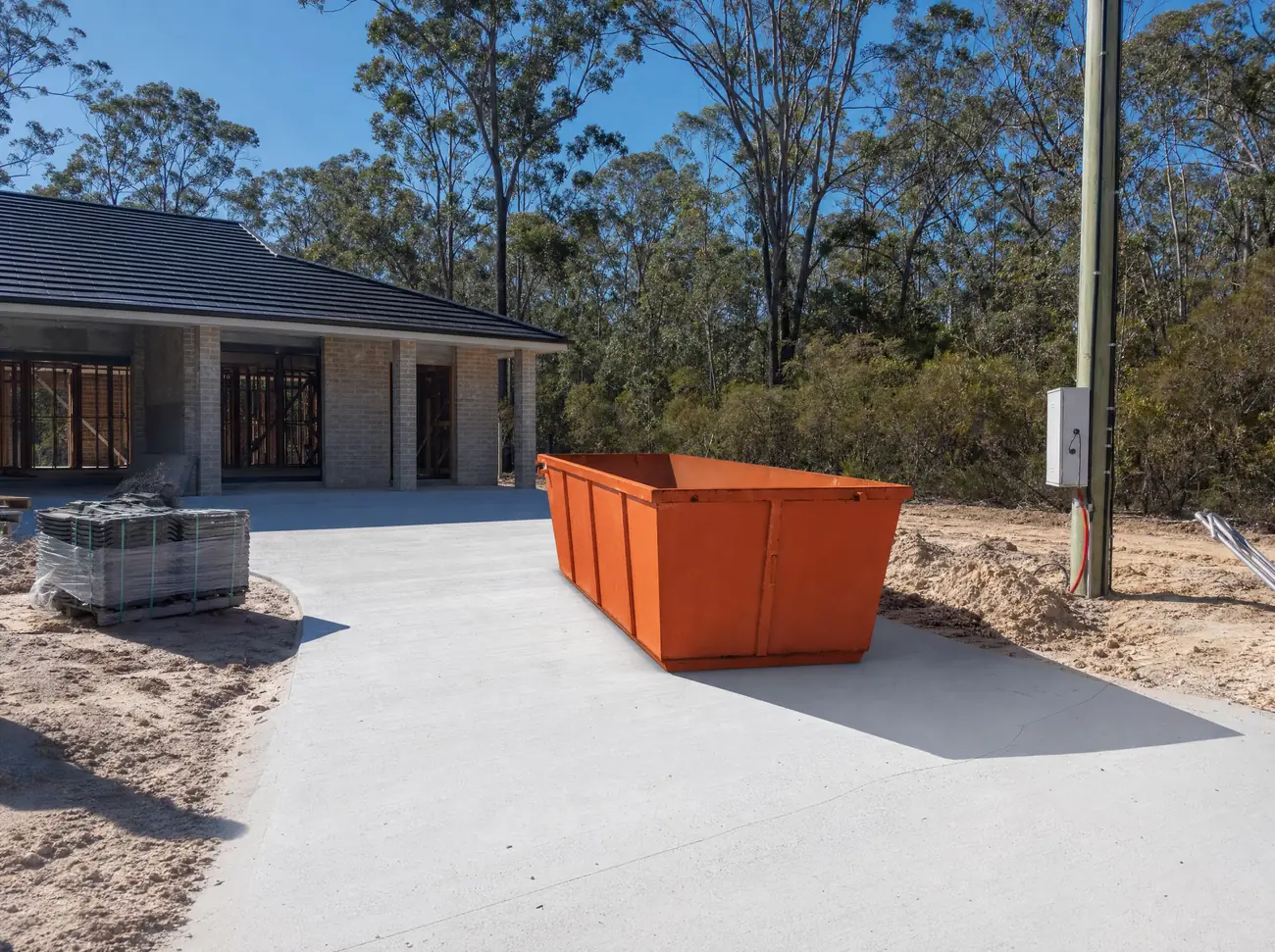 An empty orange skip bin sitting lengthways on the new concrete driveway of a partly built house on a large Medowie block, sandy soil and stacked roof tiles nearby, scribbly gums and banksia at the boundary