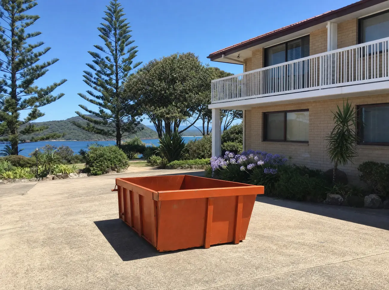 An empty orange skip bin sitting lengthways on the concrete driveway of a low-rise brick unit block at Nelson Bay, Norfolk Island pines and coastal shrubs around it and a glimpse of the bay behind