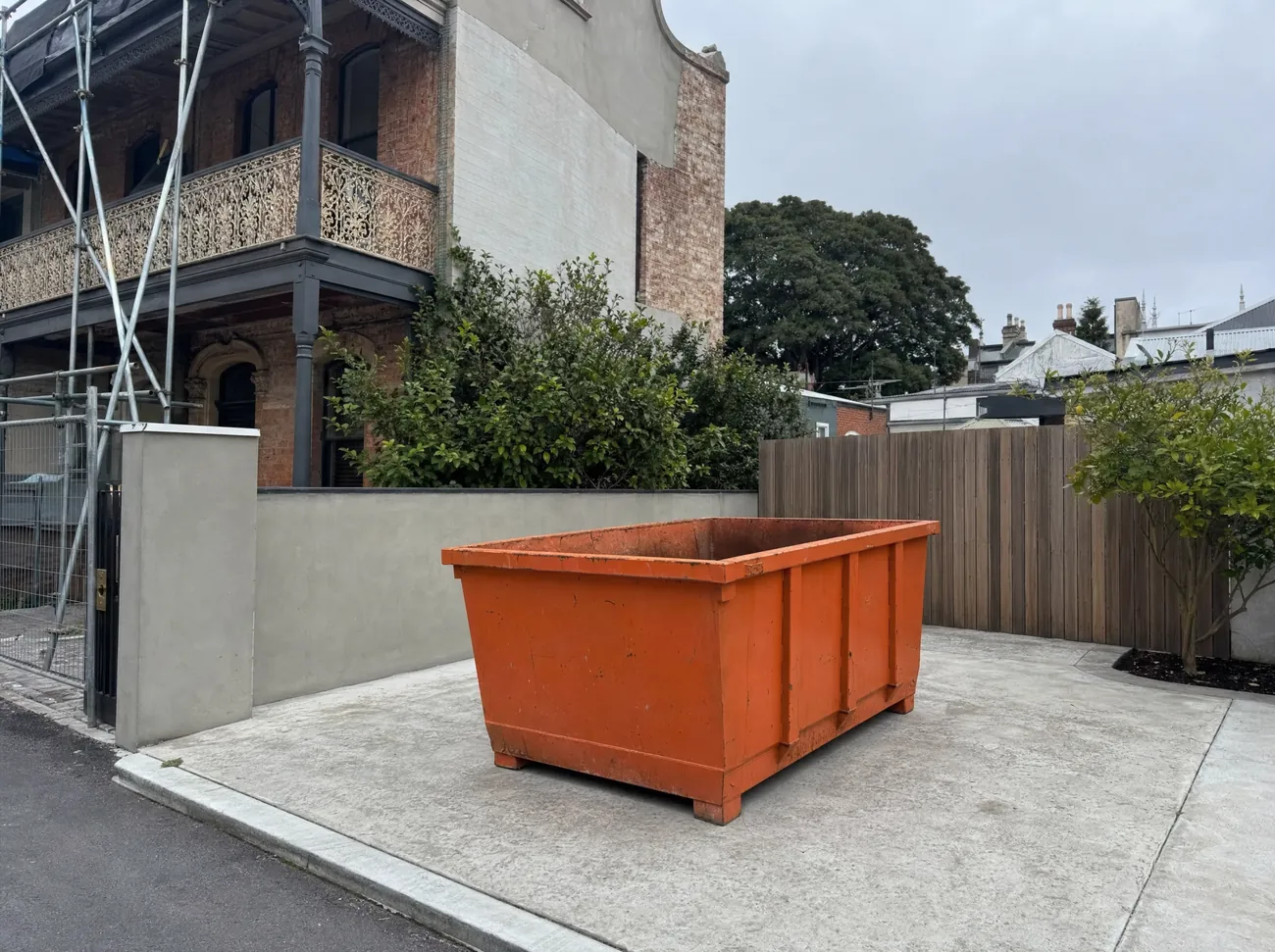 An empty orange skip bin sitting lengthways on the concrete hardstand beside a two-storey Newcastle terrace mid-renovation, iron lace balcony and scaffolding above, a Port Jackson fig on the street and inner-city rooftops behind
