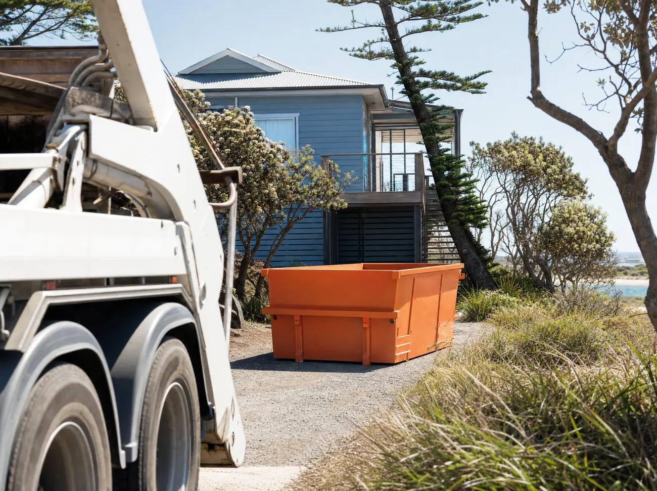 An empty orange skip bin sitting lengthways on the driveway of a raised weatherboard beach house at Port Stephens, a skip truck in the foreground, coastal banksia, a Norfolk pine and a sandy verge around it