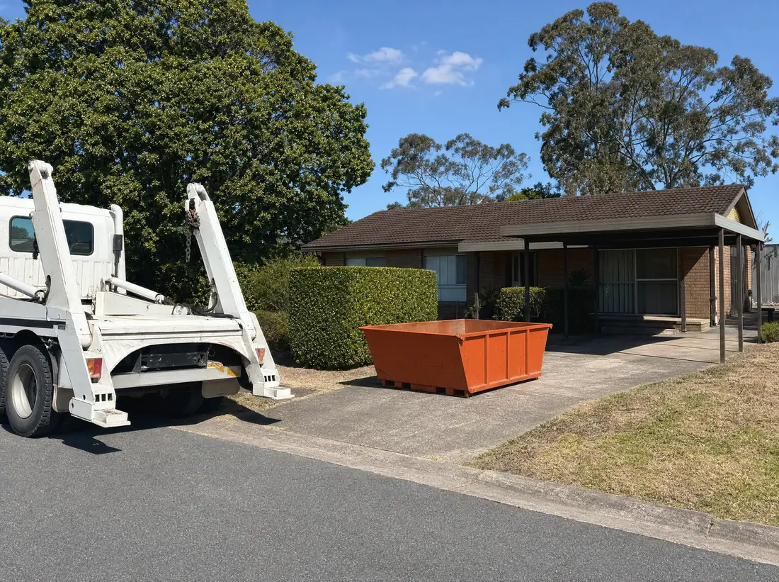 An empty orange skip bin sitting lengthways on the concrete driveway of a 1970s brick-and-tile home in Raymond Terrace, a skip truck parked on the street and tall gums behind the roofline