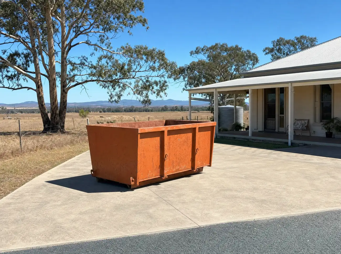 An empty orange skip bin sitting lengthways on the wide concrete driveway of a verandahed homestead-style house near Singleton, a rainwater tank alongside, huge river red gums and open dry paddocks behind