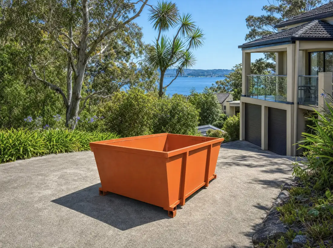 An empty orange skip bin sitting lengthways on the sloping driveway of a two-storey rendered home at Warners Bay, a cabbage-tree palm and gums in the front garden and the lake visible in the distance
