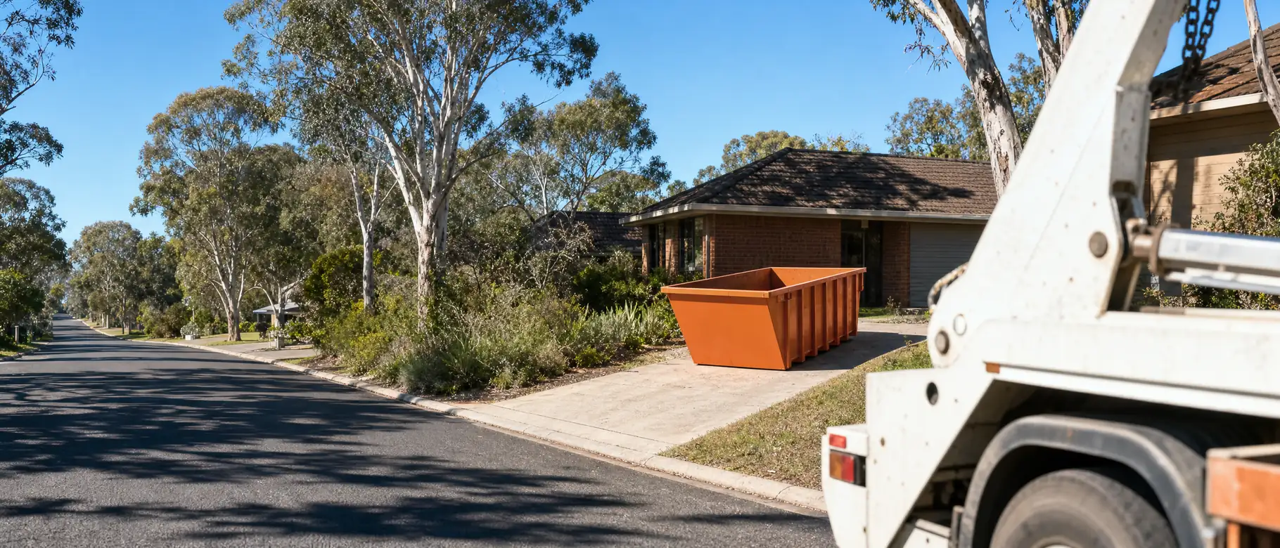 An empty orange skip bin sitting lengthways on a suburban driveway in the Hunter, a skip truck in the foreground, bright hard Australian sunlight