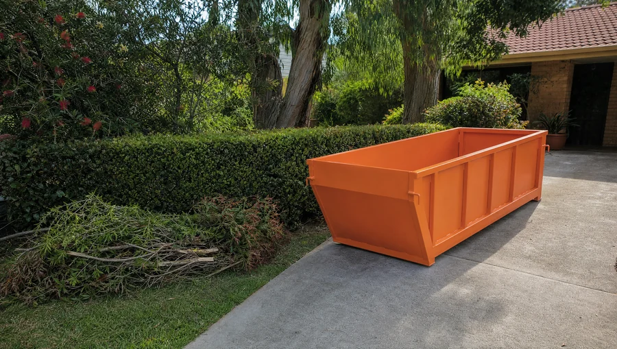 An empty orange skip bin sitting lengthways on a Hunter driveway beside an established garden, a freshly pruned hedge and a pile of cut branches on the lawn alongside